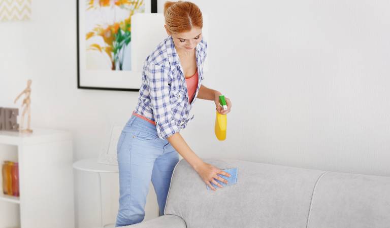 Woman in check shirt and jeans scrubbing sofa with a cloth and holding a bottle in her hand