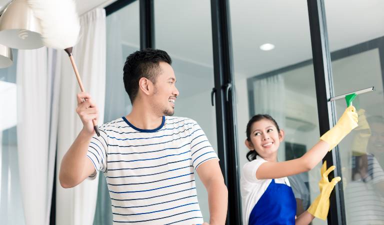 Man holding a duster and woman holding a bottle in hand seeing each other and smiling.