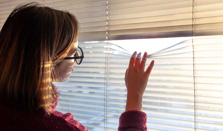 Woman in spectacles is seeing outside from window blinds