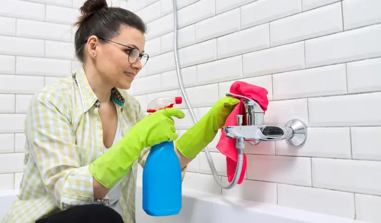 Woman in green gloves holding a blue bottle cleaning water pipe with a red cloth inside a bathroom.