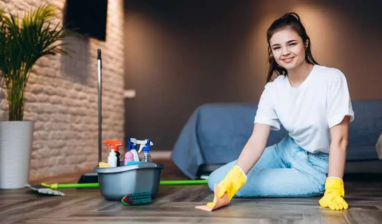 A lady in white top, blue jeans and yellow gloves sitting on floor with a cleaning basket.