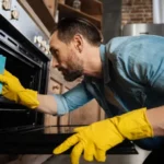Appealing bearded male cleaner leaning over oven while cleaning it and wearing gloves