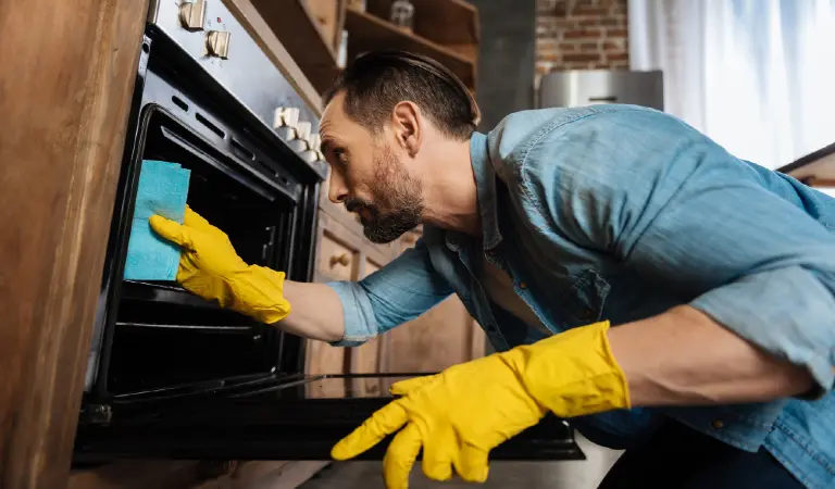 Appealing bearded male cleaner leaning over oven while cleaning it and wearing gloves